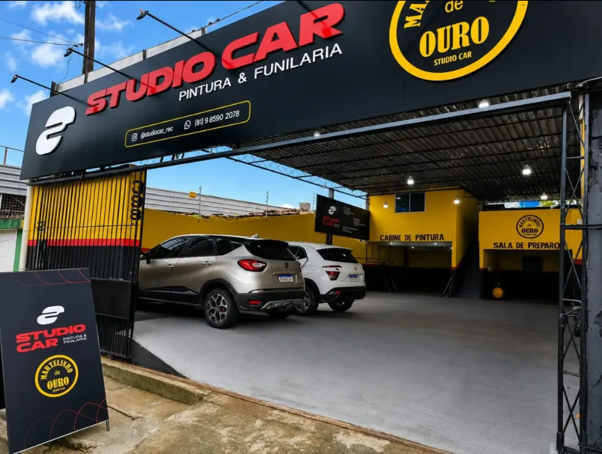 Fachada da StudioCar Pintura & Funilaria, com letreiro preto e vermelho, paredes amarelas e carros na oficina.